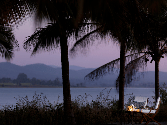 Two chairs and a table kept near the lakeside with coconut trees next to the chairs and faraway hills in view during twilight hour at The Serai Kabini - the best resort in Kabini.