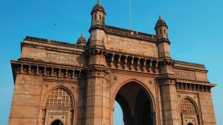 A view from below of the magnificent Gateway of India with intricate carvings on it and a blue sky in the background.