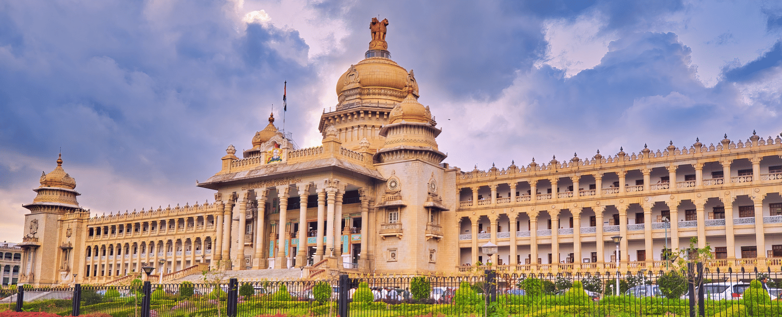 Vidhana Soudha building in Bengaluru under a cloudy sky, featuring ornate architecture and a neatly manicured lawn