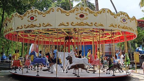 A colourful carousel with children riding ornate horse figures spins joyfully in a park under the shade of trees.