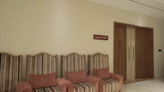 A Corridor of Pride Resort, Rajkot light walls, a small seating area with striped benches, and a wooden door, providing a simple yet functional space