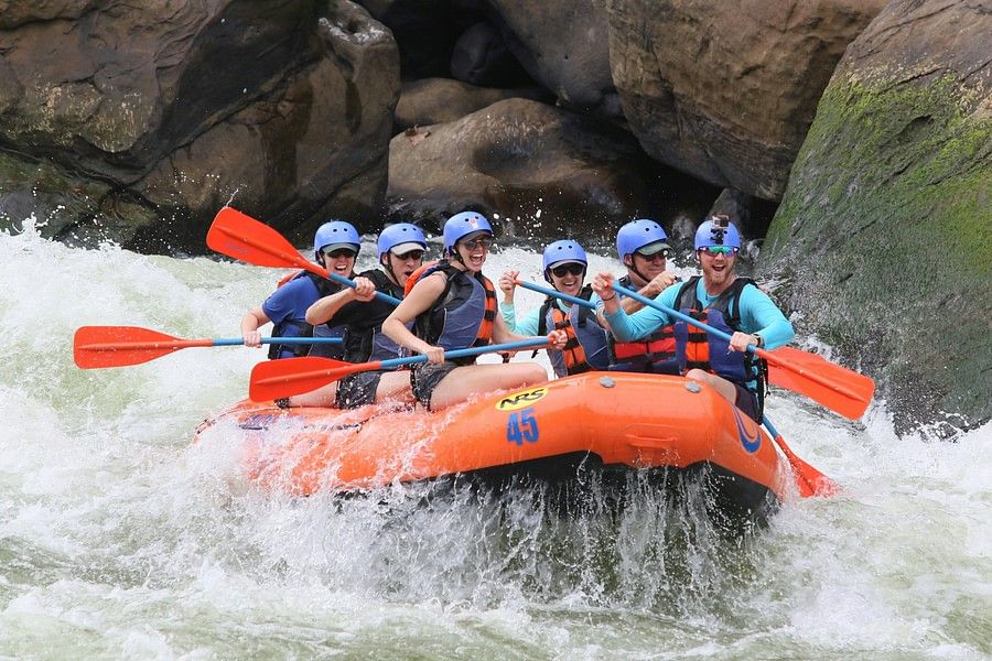 alt-text Another group of people in blue life jackets and helmets navigating through white water rapids in an orange raft, with large rocks and turbulent water around.