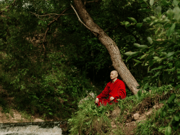 A monk in red robes sitting peacefully by a river under a green tree.