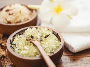 A bowl of yellow Himalayan salt scrub with dried herbs, next to a white towel and a flower.