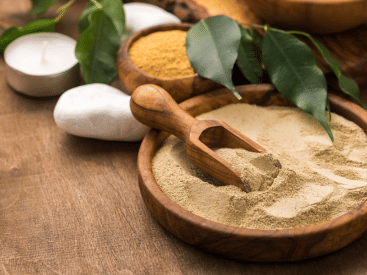 A bowl of beige body scrub powder with a wooden scoop, flanked by leaves, a candle, and a massage tool.