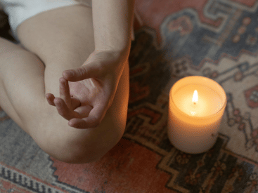 A close-up of a meditative hand gesture next to a lit candle on a patterned rug.
