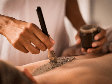 A person receiving a body scrub treatment, with a close-up of hands applying the scrub on their back using a brush.