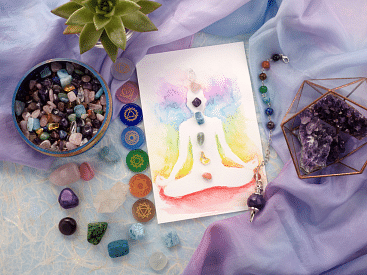 A colorful illustration of a meditating figure with chakras highlighted, surrounded by crystals, gemstones, and a succulent plant on a pastel fabric background.