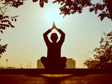 A silhouette of a person sitting cross-legged with hands in a prayer position above their head, meditating outdoors at sunrise or sunset.