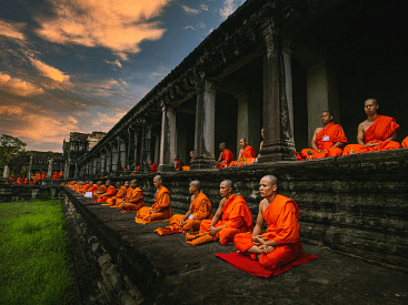 A group of Buddhist monks in orange robes meditating in a long row on stone steps, with an ancient temple and a dramatic sunset sky in the background.