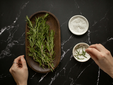 Fresh rosemary sprigs on a wooden tray with a small bowl of salt, on a dark marble countertop.