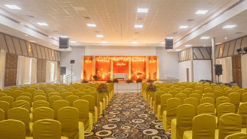 An image of a banquet hall with several chairs, speakers, cameras, and a stage set up on the front part of the hall- The Renai Cochin