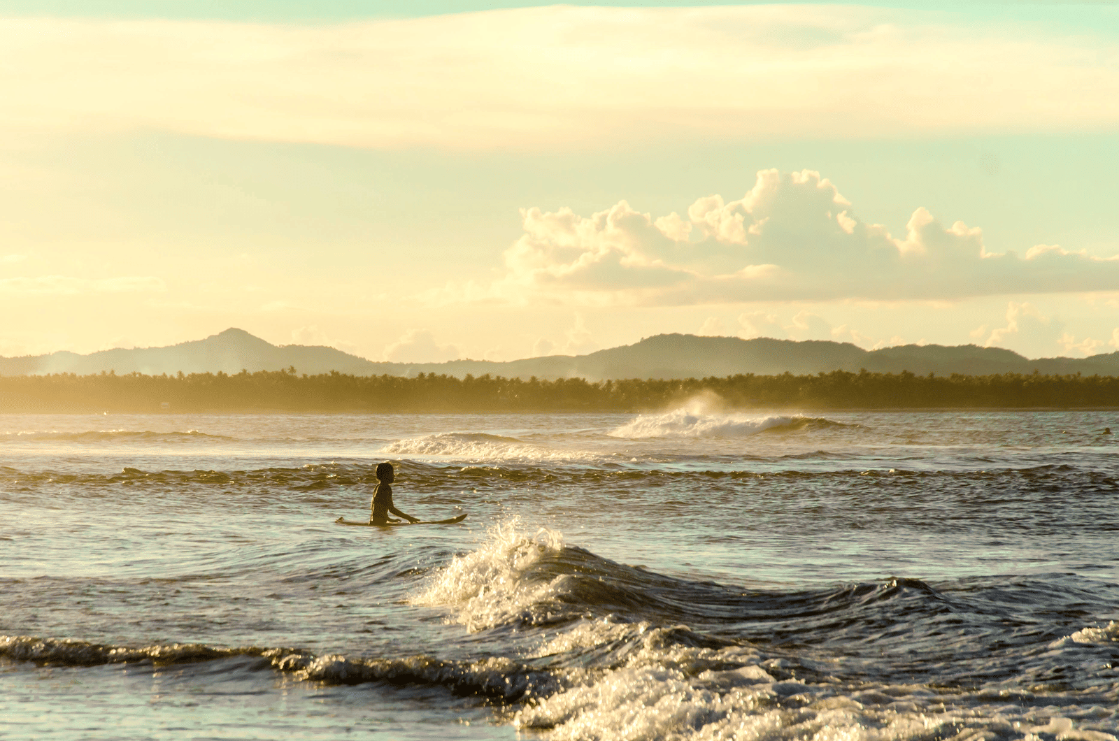 A lone surfer sitting on a board in golden-hour waves off Siargao, with palm-lined shoreline and hills on the horizon