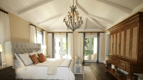 A large bedroom with high ceilings features a upholstered king size bed, chandelier, wooden wardrobe, and french doors.