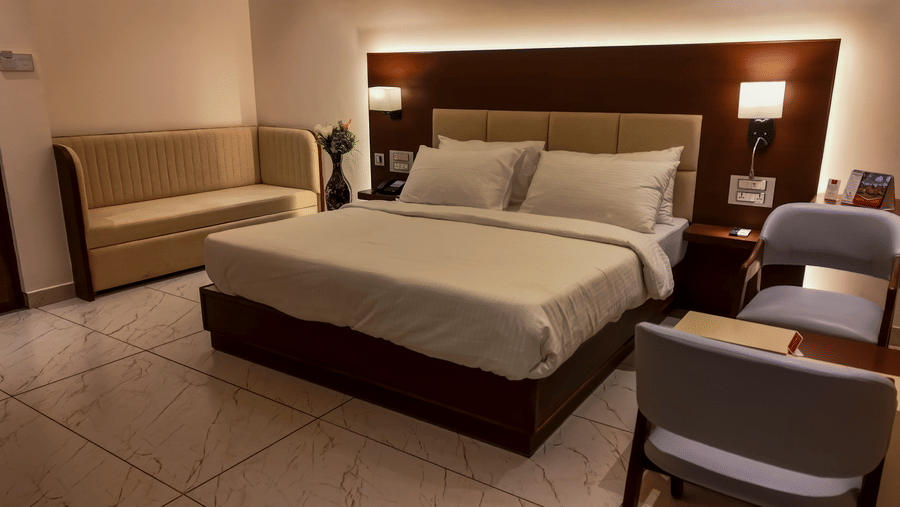 A hotel bedroom featuring a double bed with white sheets, a sofa, a side table with chairs, and wall-mounted lamps | Lords Sahyadri Resort, Shivamogga