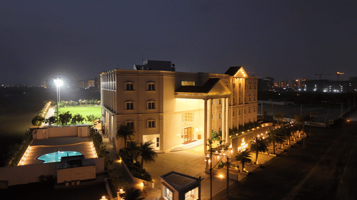 An imposing multi-story building of Pride Resort Rajkot which is a symmetrical facade, surrounded by a parking area and open land.