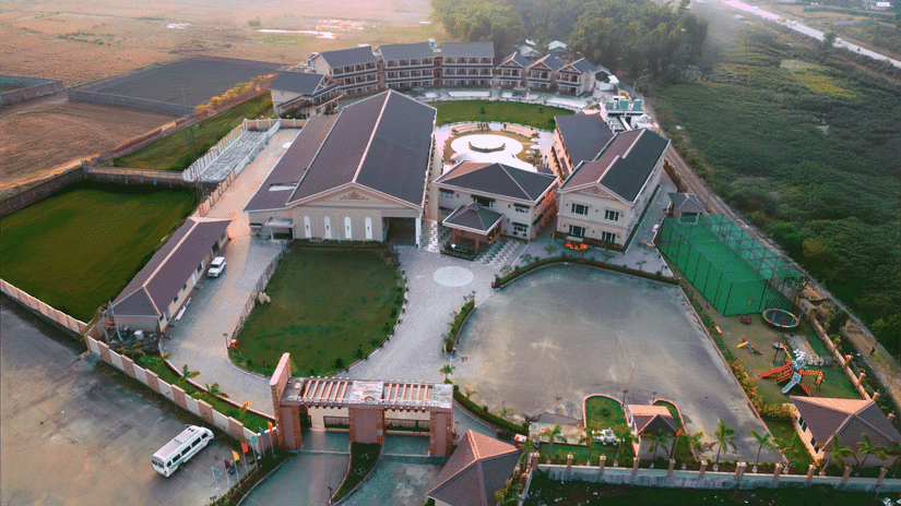 Aerial view of a large hotel or resort with multiple buildings, green lawns, a playground, and a sports court, surrounded by open fields. 