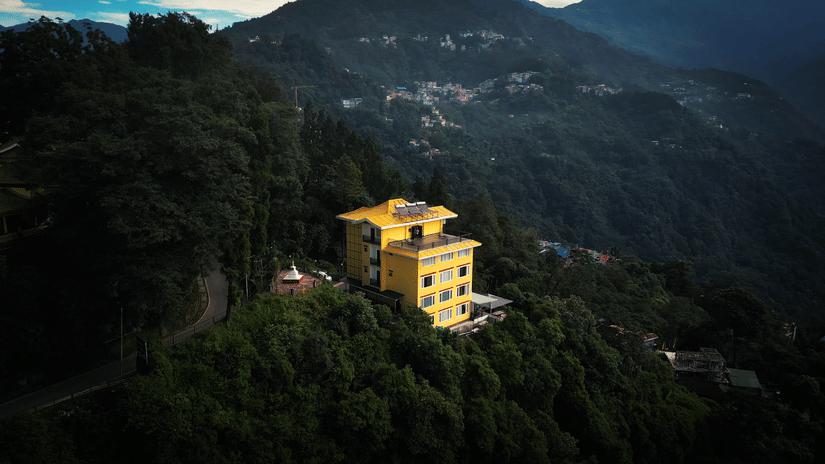 Aerial view of the yellow hotel building perched on a lush green hillside with a scenic valley backdrop at Voyage The Royal Retreat.