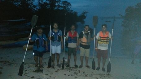 Five friends stand on a beach at dusk, holding paddles, wearing life jackets, and ready for an early morning kayaking trip.