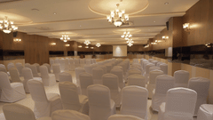 A spacious, well-lit conference or event hall at Pride Resort, Rajkot completely furnished with numerous rows of white chairs, awaiting attendees for a large gathering