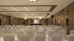 A large, brightly lit conference hall at Pride Resort, Rajkot showcasing numerous rows of neatly arranged white chairs facing a central stage or podium