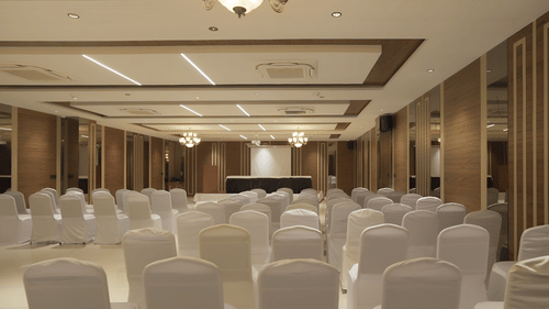 A large, brightly lit conference hall at Pride Resort, Rajkot showcasing numerous rows of neatly arranged white chairs facing a central stage or podium