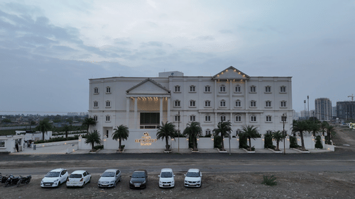 An imposing multi-story building, of Pride Resort Rajkot, featuring a symmetrical facade, surrounded by a parking area and open land.