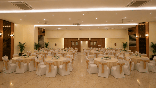 Lavender banquet hall at Tulip Inn, Hoodi Whitefield, Bengaluru, set up with round tables, chairs, covers, ties, and indoor potted plants.