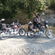 2 motorcyclists with loaded bikes taking a break along a gravel path near a shallow stream crossing.