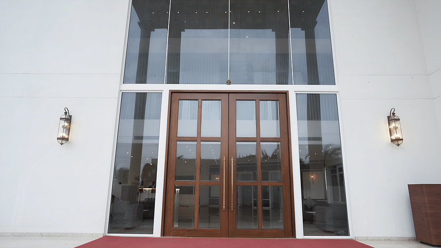 The grand entrance of Pride Resort, Rajkot featuring large wooden doors flanked by tall glass panels and classic outdoor wall sconces, welcoming visitors