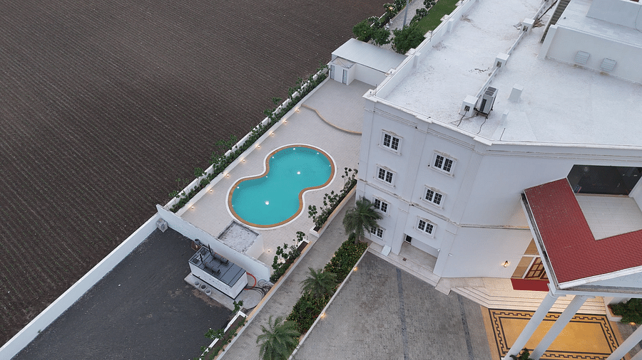 Aerial view of a green, figure-8 shaped swimming pool surrounded by light grey tiling and sparse trees - Pride Resort Rajkot