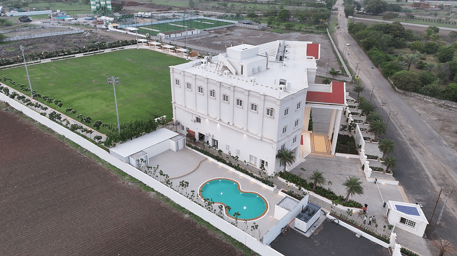 A multi-story white building of Pride Resort, Rajkot featuring a swimming pool, surrounded by green lawns and undeveloped land
