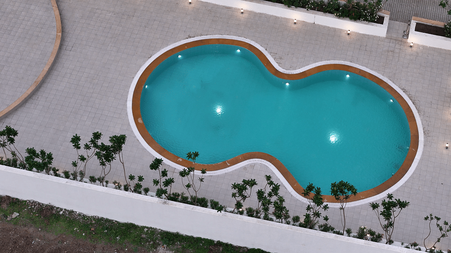 Two-lobed swimming pool at Pride Resort, Rajkot featuring a clear turquoise water, surrounded by paved areas and greenery