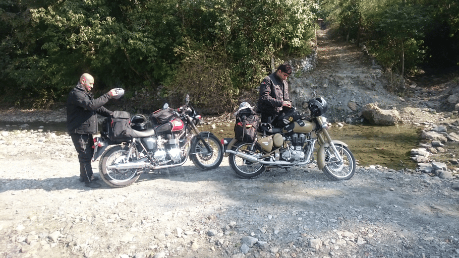 2 motorcyclists with loaded bikes taking a break along a gravel path near a shallow stream crossing.