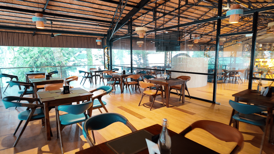 Above Clouds Bar at DNC Shevaroys Resort & Spa featuring colourful chairs, wooden tables, and large windows overlooking the forest.