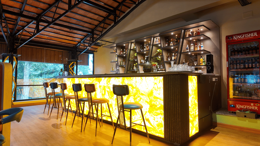 A backlit golden bar counter with tall stools and shelving for bottles at the DNC Shevaroys Resort & Spa.