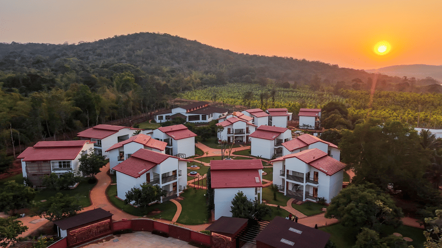 An elevated view of a cluster of white cottages with red roofs at sunset, with hills and trees in the background | Lords Sahyadri Resort, Shivamogga