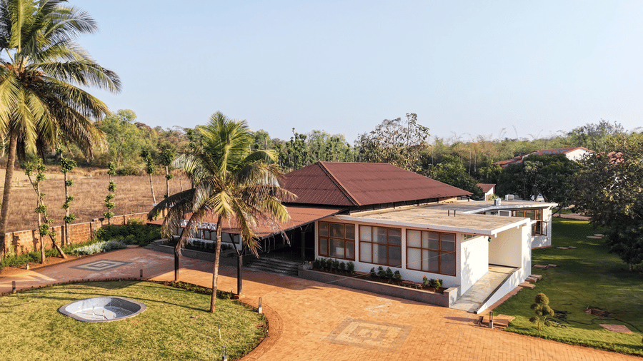 An elevated view of a resort building with a red tiled roof, a paved driveway, and palm trees on a grassy lawn | Lords Sahyadri Resort, Shivamogga