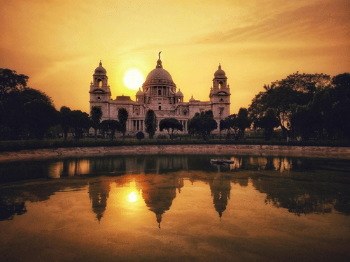 A view of Victoria Memorial, Kolkata, reflected in the water during a golden sunset with trees on either side.