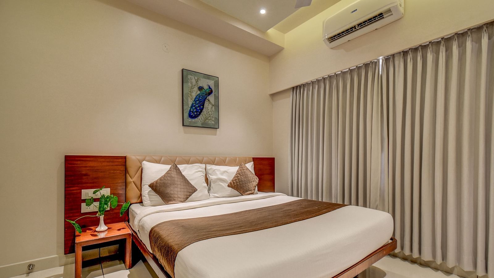 A spacious room at Harigovinda's JNStay with a large bed, a bedside table, large windows fully covered with curtains, and a wall-mounted photo frame above the headboard