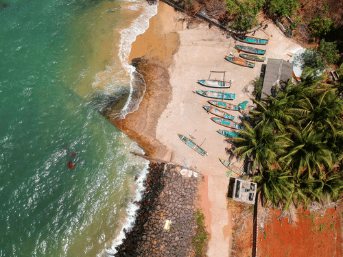 An aerial top-down view showing multiple small boats parked on a sandy beach coastline next to green palm trees and waves meeting the shore.