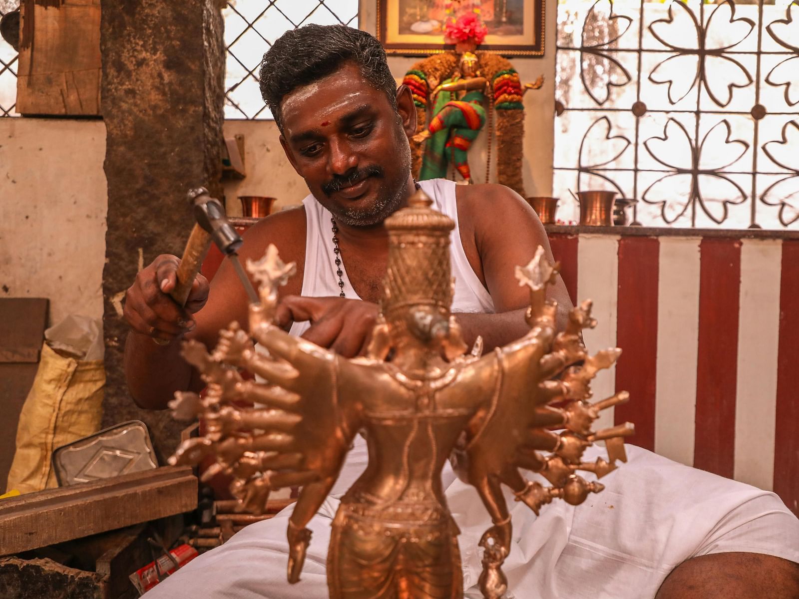 A craftsman meticulously working on a bronze idol of a multi-armed deity, using a hammer and chisel.