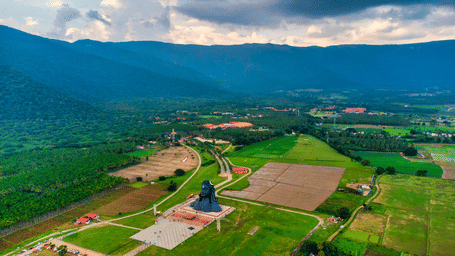 An aerial view of the large black Adiyogi Shiva statue standing in a vast green field, surrounded by lush forests and the hazy peaks of a mountain range.