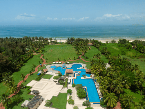 An aerial view of Kenilworth Resort & Spa, Goa, with vast blue ocean at the far end and a swimming pool, a lawn area, pathways and many trees surrounding the property. It is the best place for a long stay in Goa.