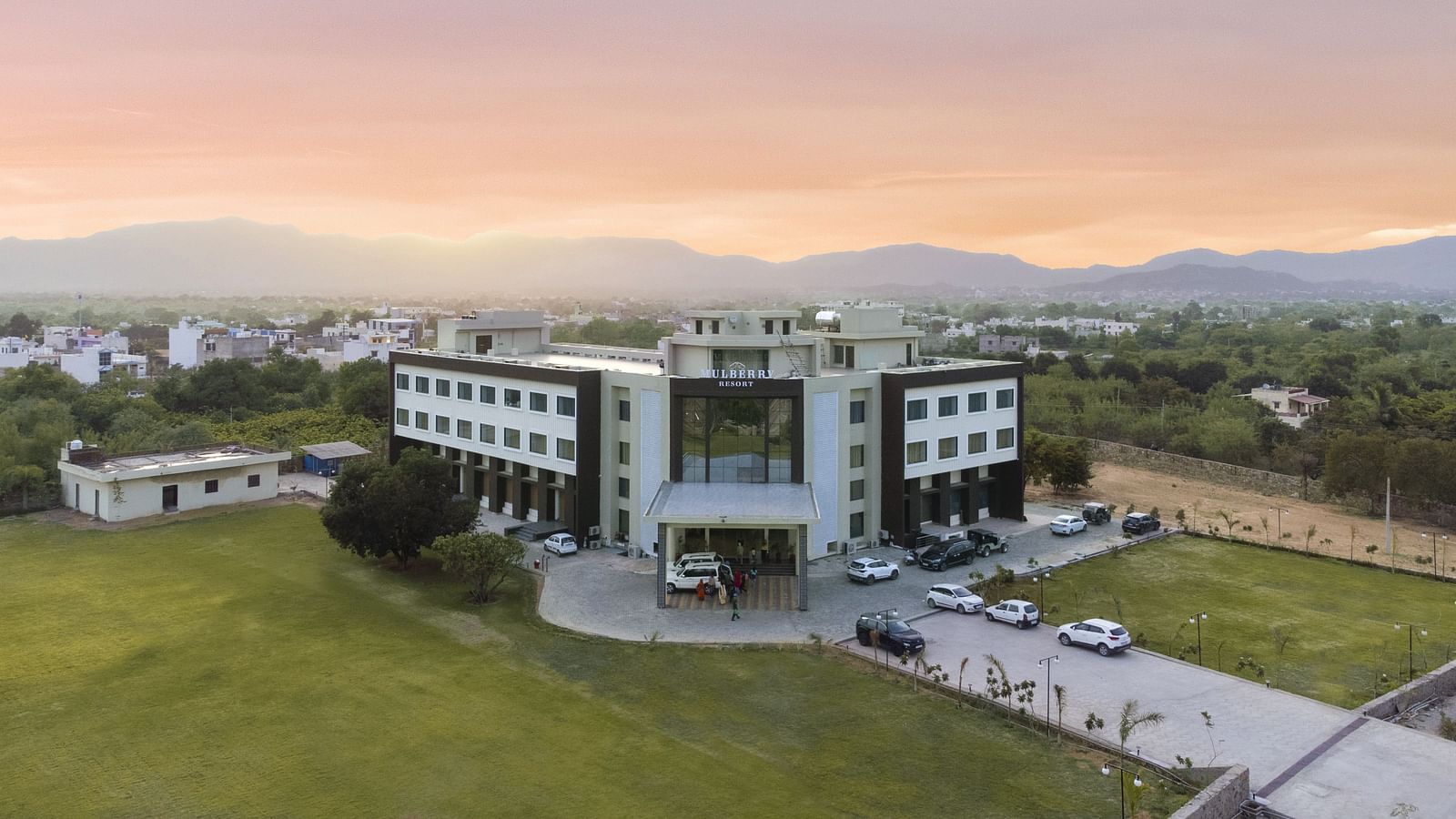 An aerial view of the Bamboo Saa Mulberry Resort, Pushkar at sunset or sunrise surrounded by green lawn and the backdrop of hills.