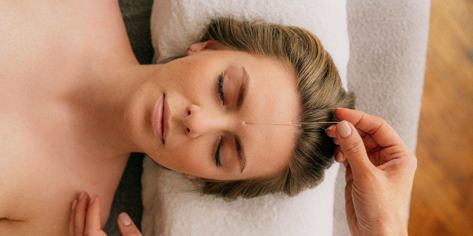 A client with her eyes closed lies on a massage table as a therapist places a thin needle for an acupuncture treatment on her forehead.