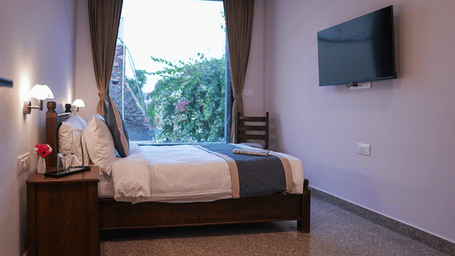 A side view of a bedroom with a wooden double bed, white linens, a blue runner and a wall-mounted TV | Daksh the Fateh Retreat