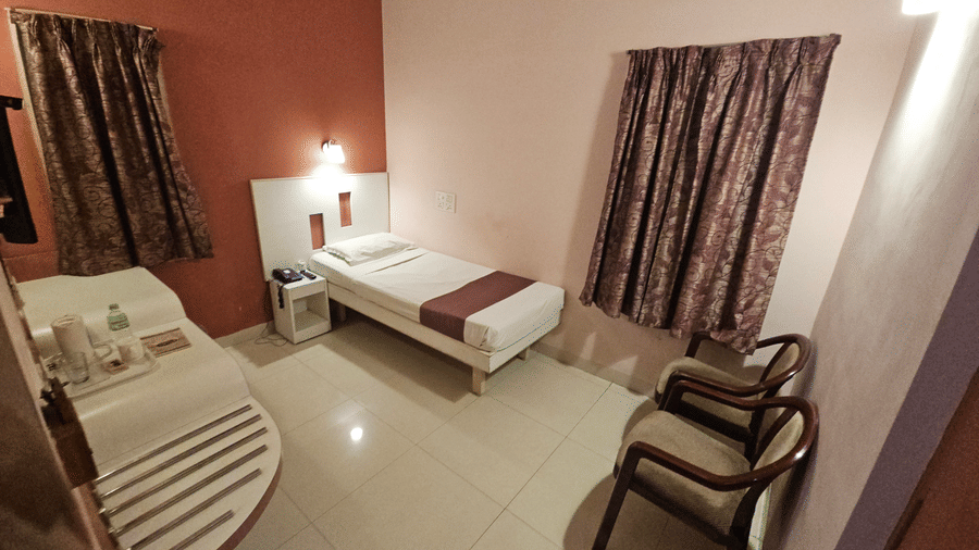 A simple hotel guest room with a single bed, bedside table, dark curtains over two windows, and 2 wooden armchairs at Hotel Pandian.