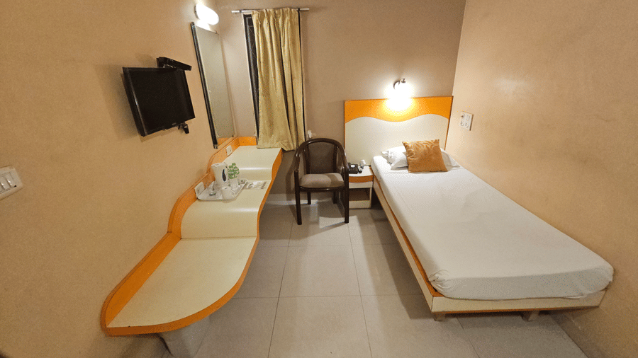 A minimalist hotel single room featuring a single bed with white linens, a wooden desk unit, and a wall-mounted TV.