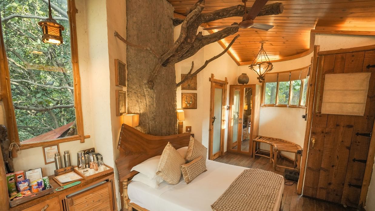 A bedroom with a bed, a lamp, and warm lighting, interior setting with wooden elements and large windows - The Den Corbett Resort & Spa, Kumeria.